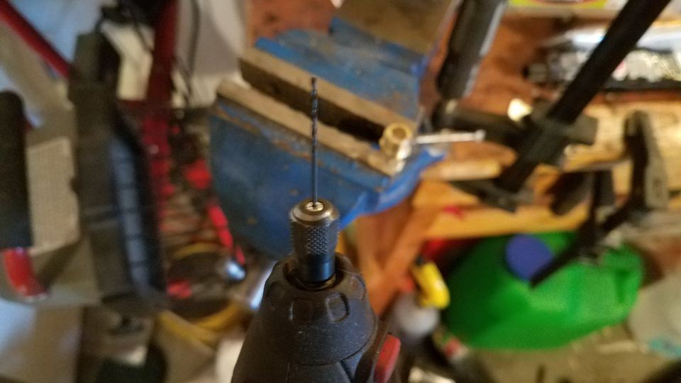 Close-up of a small drill bit held in a rotary tool chuck, with a blue vise and other tools in the background