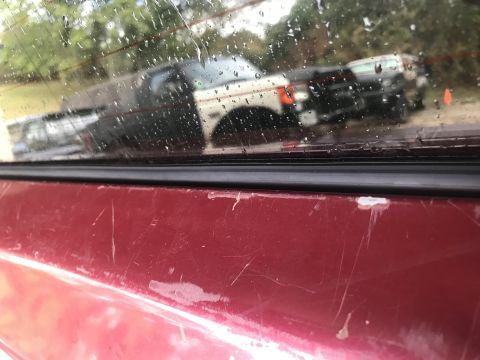 1994 Ford Bronco - Scratches mar the red paint of a vehicle, with reflections of other trucks visible in the rain-streaked window.