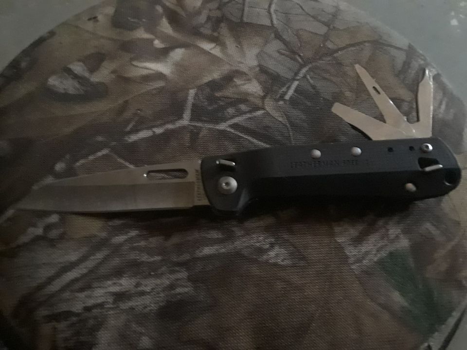 Black multi-tool knife with blade and other tools extended, resting on camouflage fabric