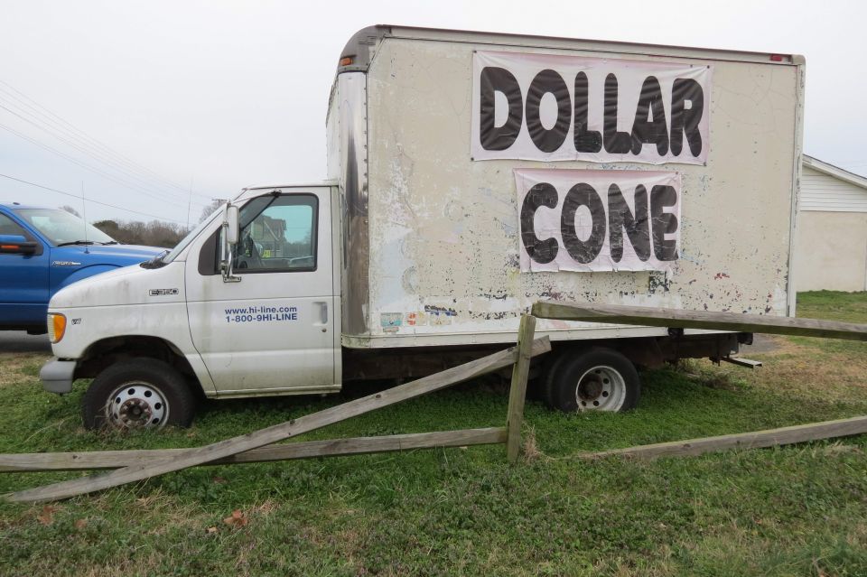 White Ford E350 box truck with 'DOLLAR CONE' signs parked behind a broken fence on grass