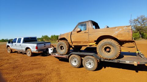 1995 Ford Bronco - A mud-caked 1995 Ford Bronco is transported on a trailer, pulled by a silver pickup truck under a clear blue sky.