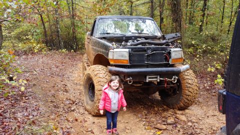 1995 Ford Bronco - A young girl stands before a heavily modified pickup truck on a wet, wooded trail.