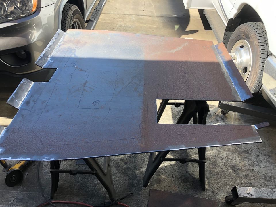Sheet metal frame part with cutouts and scribe lines, resting on sawhorses between two vehicles