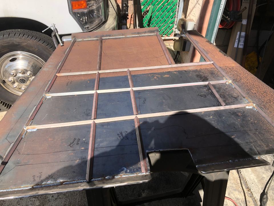 Metal frame with welded grid pattern, some scribed lines and text. A white truck wheel is visible to the left