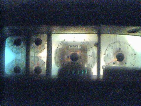 1989 Ford Bronco - Dark, grainy nighttime shot of an instrument cluster with white-faced gauges: four small round gauges at left, a large center speedometer w…