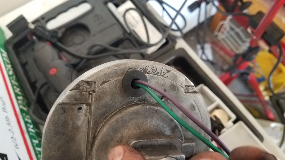Close-up of a metal part with three wires (green, purple, black) coming out of a rubber grommet. Some stamped text is visible above the grommet