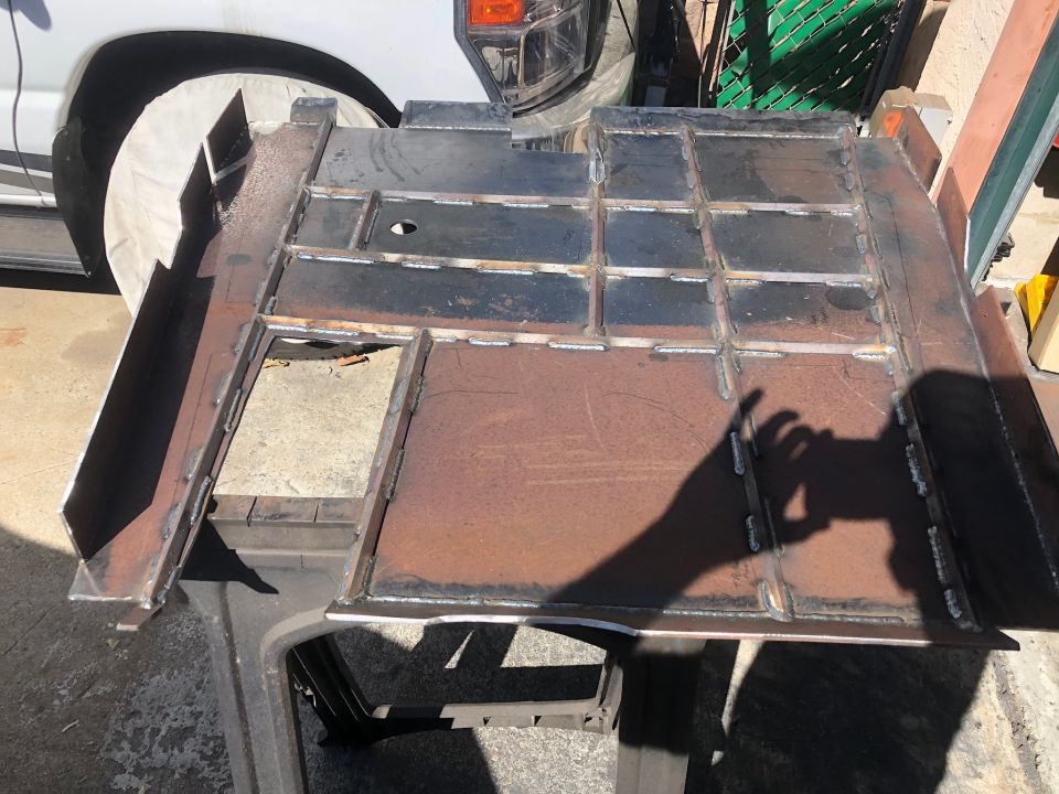 Fabricated metal frame section with visible welds, resting on a stand. A white vehicle's tire and fender are partially visible