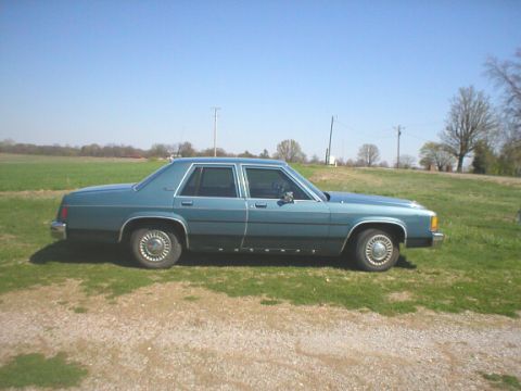 1987 Ford Crown Victoria LTD - A blue sedan sits on a gravel patch in front of a field under a clear blue sky.