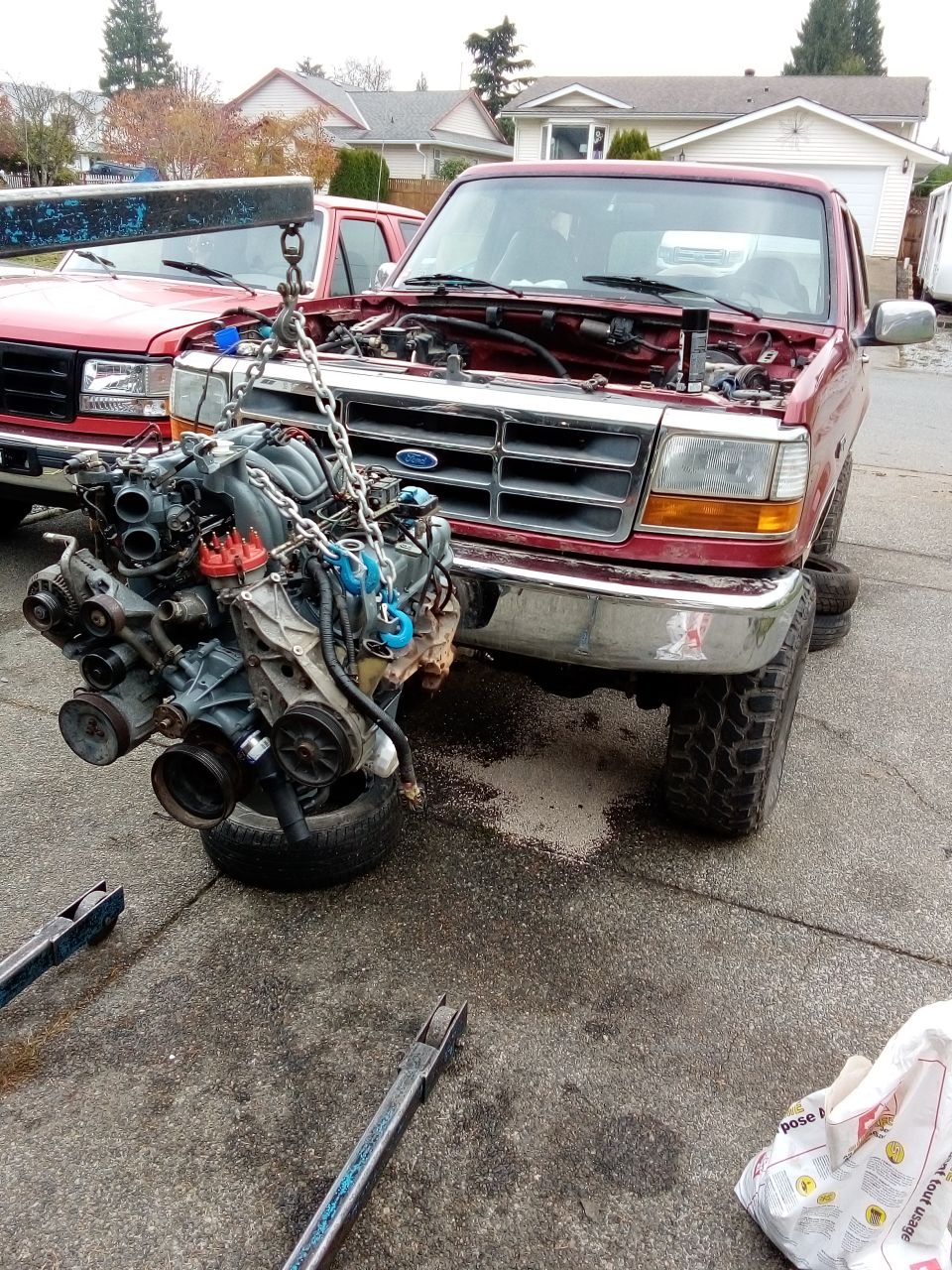 1994 Ford Bronco with hood open, engine being removed by hoist. Another red Ford truck is behind it