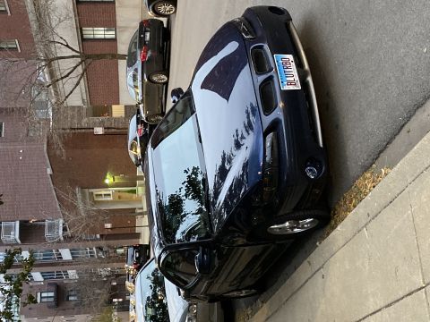 Dark blue sedan parked on street, license plate BLUTRBQ. Other cars and apartment building in background