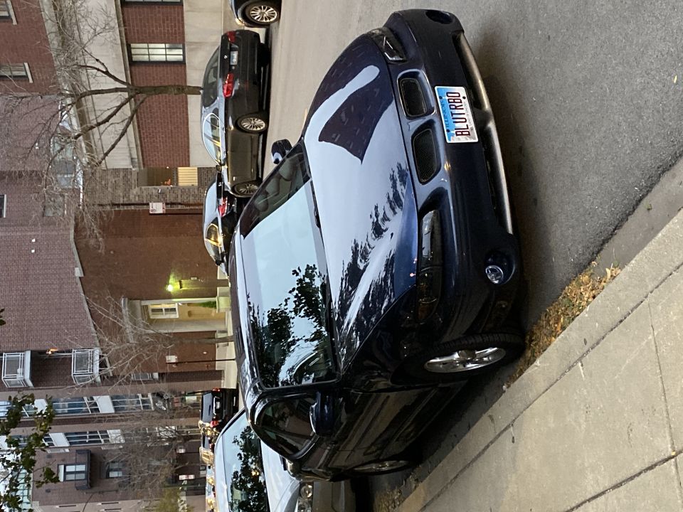 Dark blue sedan parked on street, license plate BLUTRBQ. Other cars and apartment building in background