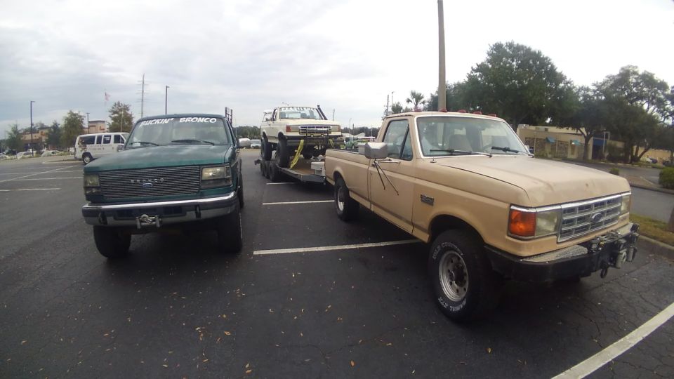 Green Ford Bronco with "FUCKIN' BRONCO" text on windshield, parked next to a tan Ford F-150 towing a white truck