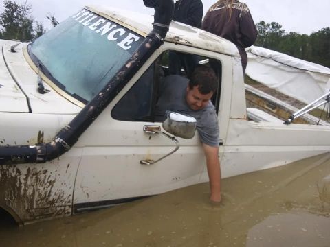 1988 Ford Bronco - A man is stuck in a white pickup truck submerged in muddy water, with a snorkel visible on the side.