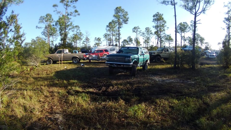 Green Bronco and other trucks parked in a muddy area with trees and a camper trailer