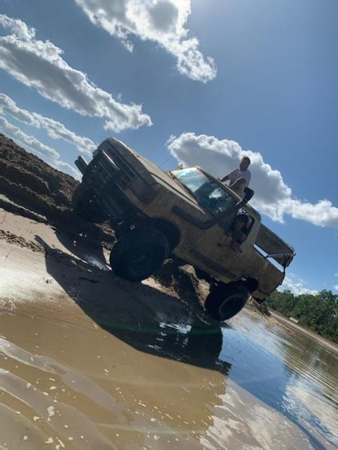 1988 Ford Bronco - A person sits on the door of a lifted pickup truck as it navigates a muddy incline with water at the bottom.