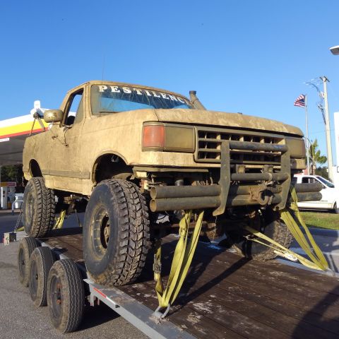 1988 Ford Bronco - This heavily modified truck, covered in mud, is secured to a trailer at a gas station.