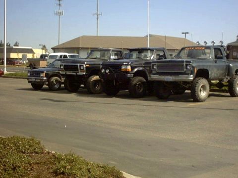 1995 Ford Bronco - Several modified trucks are parked in a lot under a clear sky.