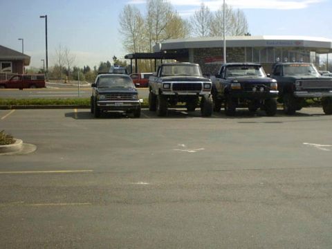 1995 Ford Bronco - Several lifted trucks are parked in a lot, with a building in the background. The middle truck is a dark color and appears to be from the e…