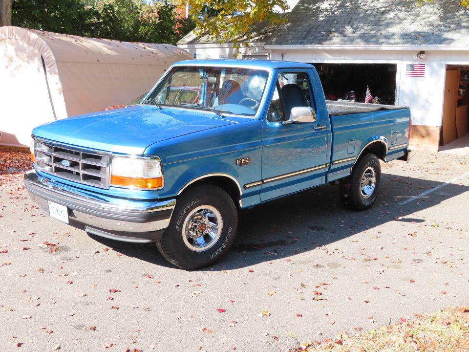 Blue Ford F-150 regular cab pickup, front three-quarter view, parked by a garage with an American flag