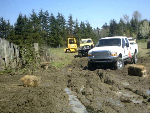 1995 Ford Bronco - A white pickup truck navigates a muddy track with other vehicles and a yellow forklift in the background.
