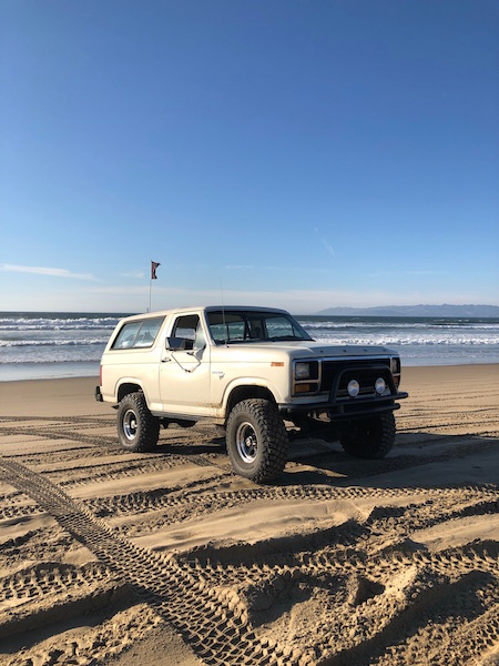 1981 Ford Bronco - A classic Ford Bronco sits on the sand near the ocean waves under a clear blue sky.