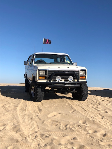 1981 Ford Bronco - A white 1981 Ford Bronco is parked on a sandy dune under a clear blue sky, flying a pink ribbon flag.
