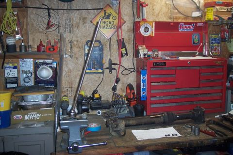 1995 Ford Bronco - A driveshaft assembly is secured in a vise on a cluttered workbench, surrounded by tools and automotive parts.