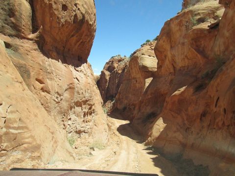1992 Ford Bronco - The vehicle is navigating a dusty trail between towering sandstone formations under a clear blue sky.