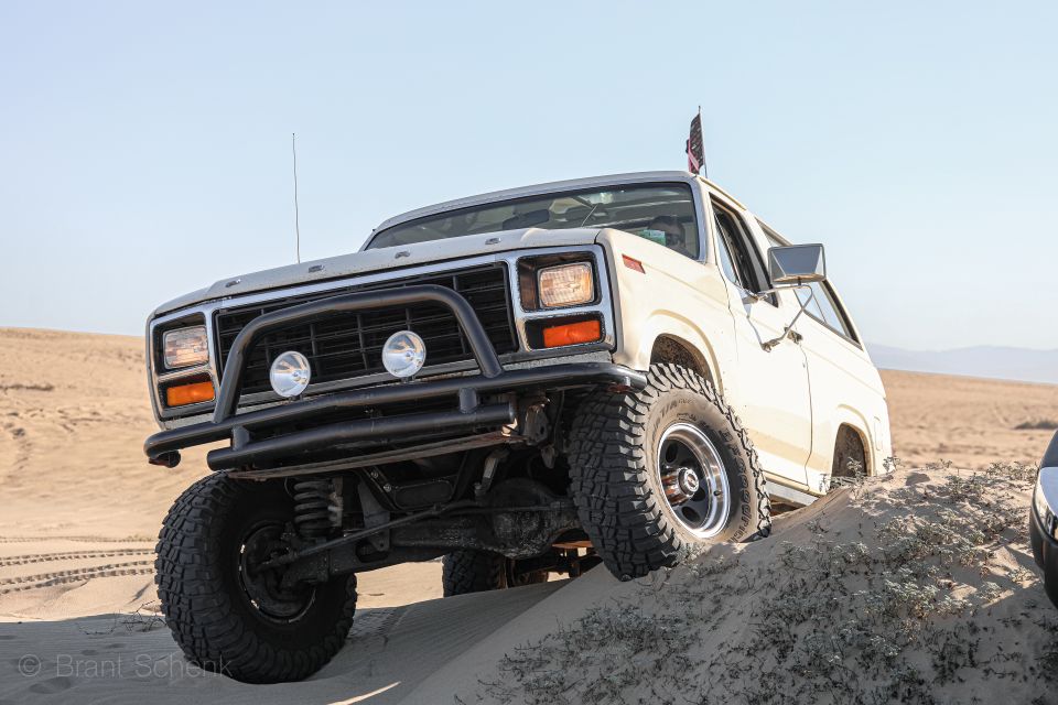 White Ford Bronco with bull bar and off-road tires driving on sand dunes. Another vehicle is partially visible on the right