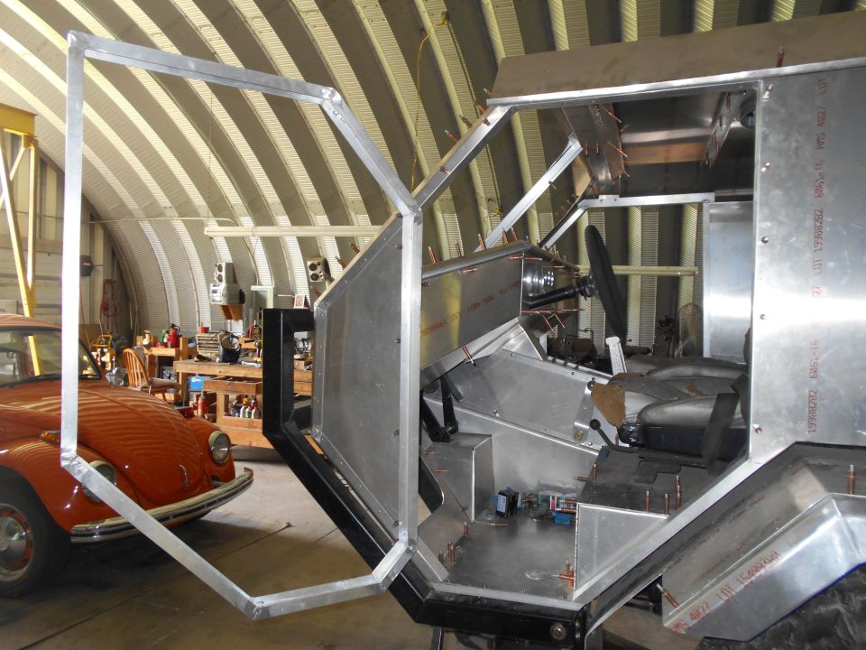 Custom vehicle interior with exposed metal panels, seats, and steering wheel. An orange car is visible to the left