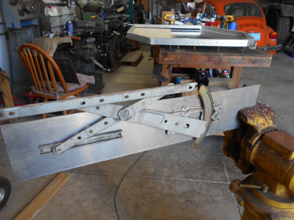 Window regulator mechanism clamped in a vise, with a wooden chair and orange car in the background