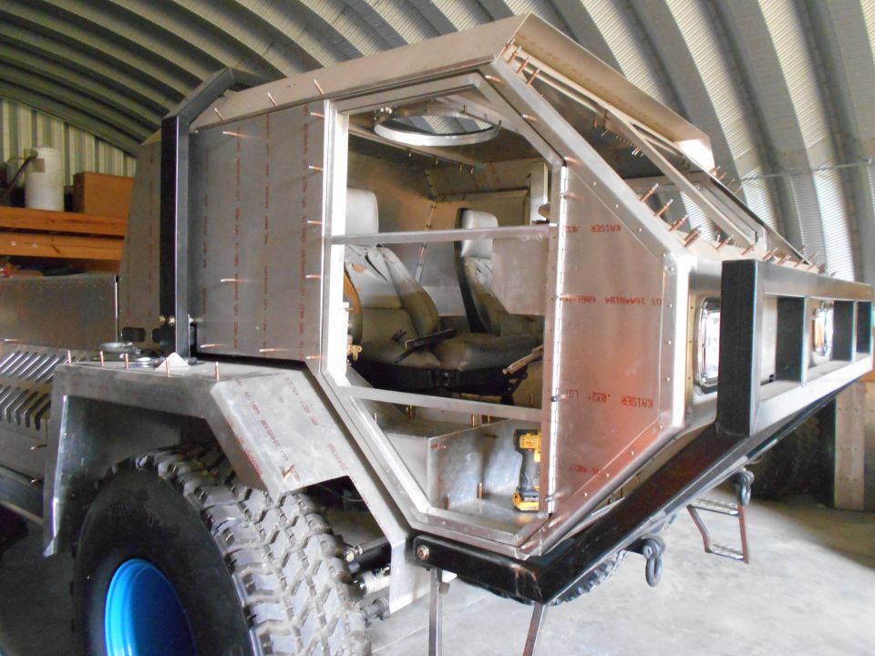 Unfinished custom vehicle with metal body panels and exposed interior, showing seats and a drill. It has large off-road tires