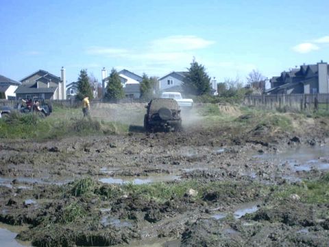 1995 Ford Bronco - An off-road vehicle navigates a muddy track, with another vehicle and houses in the distance.