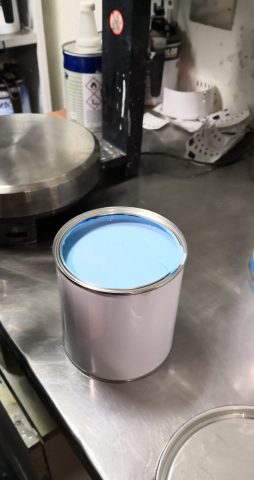 Can of light blue paint on a metal surface, with other cans and supplies in the background