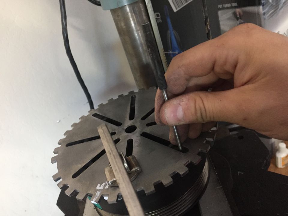 Hand using a scribe on a large toothed metal disc, possibly part of a drill press