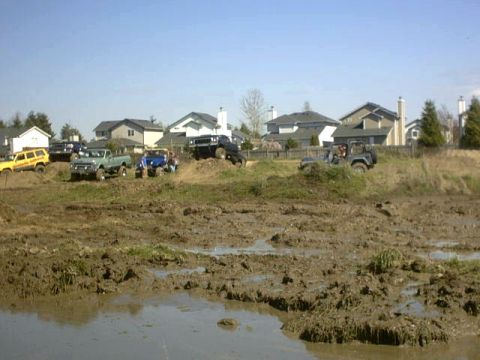1995 Ford Bronco - A collection of off-road vehicles tackle a challenging muddy course, with residential buildings visible in the distance.
