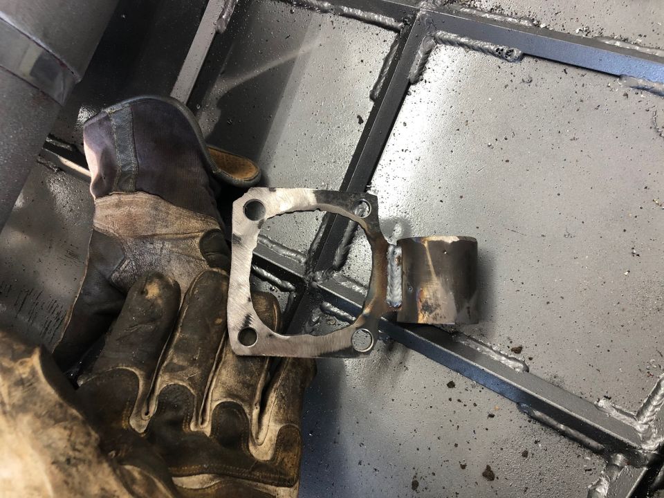 welder holding a metal flange with a cylindrical piece welded to it, over a metal grid surface