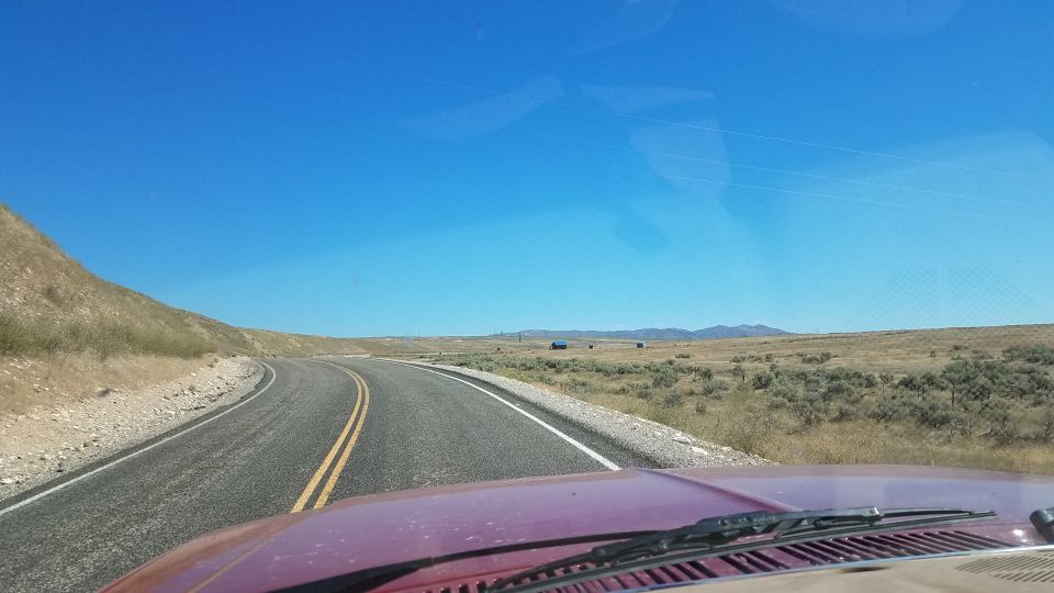 View from inside a vehicle looking out at a winding road through a dry, brush-covered landscape under a clear blue sky. A watermark reads SUPERMOTORS.NET
