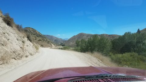 1985 Ford Bronco - View from inside a red vehicle driving on a dirt road through a scenic mountain landscape under a clear blue sky.