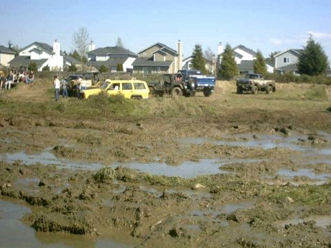 1995 Ford Bronco - Multiple off-road vehicles are seen navigating a muddy terrain, with residential buildings visible in the distance.