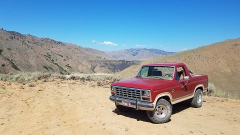 1985 Ford Bronco - A red pickup truck sits on a dusty trail overlooking a vast, arid landscape under a clear blue sky.