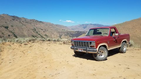 1985 Ford Bronco - A red Ford Bronco sits on a dusty trail overlooking a canyon under a clear blue sky.