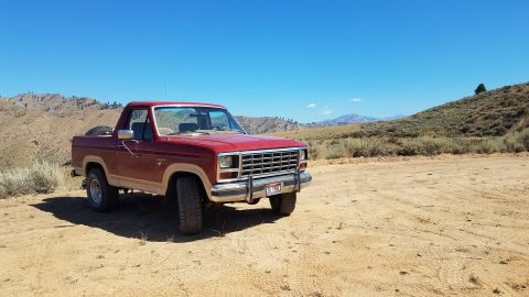 1985 Ford Bronco - This red Ford Bronco is parked on a dusty road with arid hills in the distance under a clear blue sky.