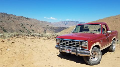 1985 Ford Bronco - A red pickup truck sits on a dusty road, with a vast, arid landscape stretching out behind it under a clear blue sky.