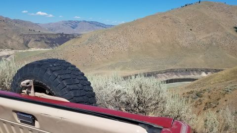 1985 Ford Bronco - The rear of an SUV with a spare tire is parked in a dry, hilly area with a small reservoir in the distance.