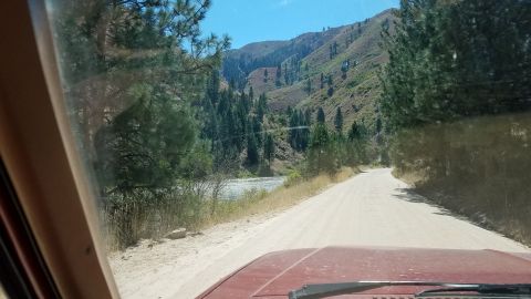 1985 Ford Bronco - Driving down a dusty road beside a river with forested mountains ahead.