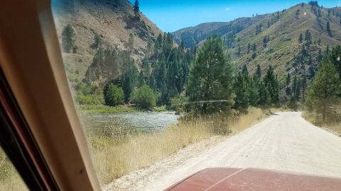 1985 Ford Bronco - A view from inside a vehicle shows a winding gravel road next to a river, with forested mountains in the background.