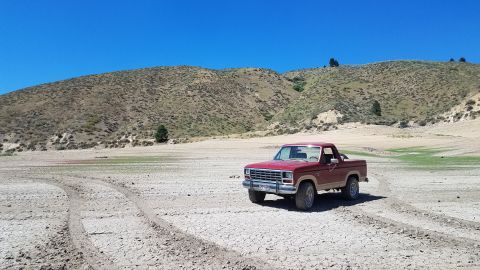 1985 Ford Bronco - A red pickup truck drives through a dry, barren landscape under a clear blue sky.