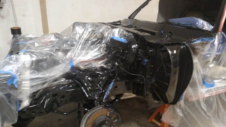 Car chassis covered in plastic sheeting, being prepped for paint. Some parts are black, others are still bare metal