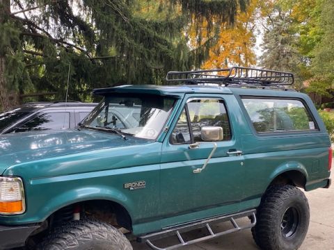 1996 Ford Bronco - This 1996 Ford Bronco is green and features a roof rack, with another vehicle visible in the background.